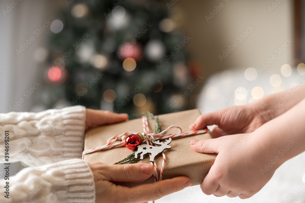 Obraz premium Woman in sweater giving a wrapped Christmas gift box to child. Glowing snow bokeh, fir tree. Winter holidays.