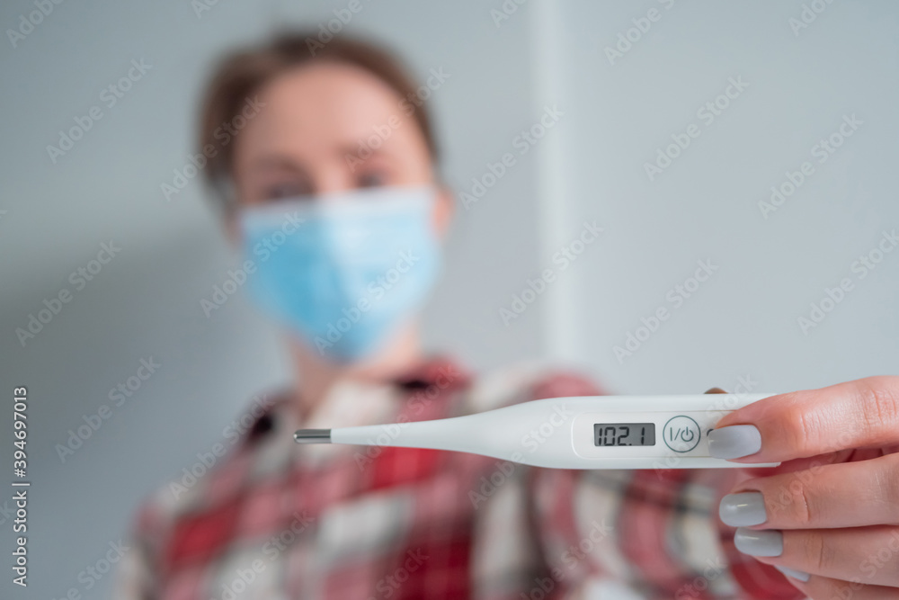 Portrait of woman in medical face mask showing digital medical thermometer with high temperature - selective focus. Healthcare, measurement, disease, covid 19, infection, coronavirus concept Portrait of woman in medical face mask showing digital medical thermometer with high temperature - selective focus. Healthcare, measurement, disease, covid 19, infection, coronavirus concept