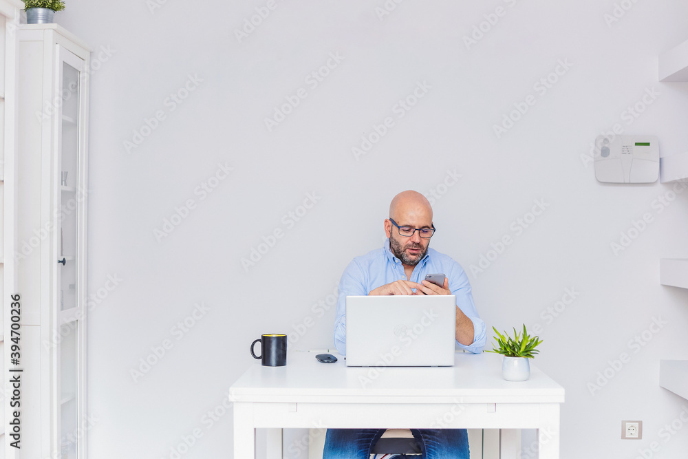 Business man teleworking from home with a laptop sending a message with his cell phone.