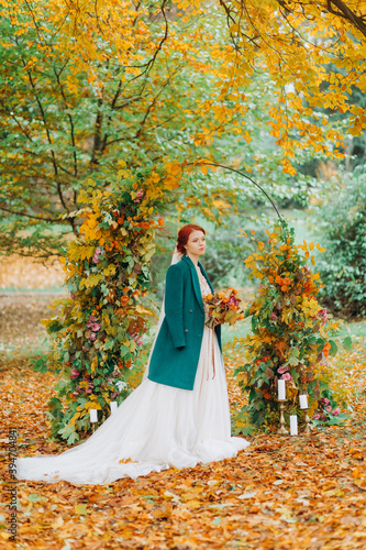 Red-haired bride in a white dress and green coat posing near the arch in Autumn Park.