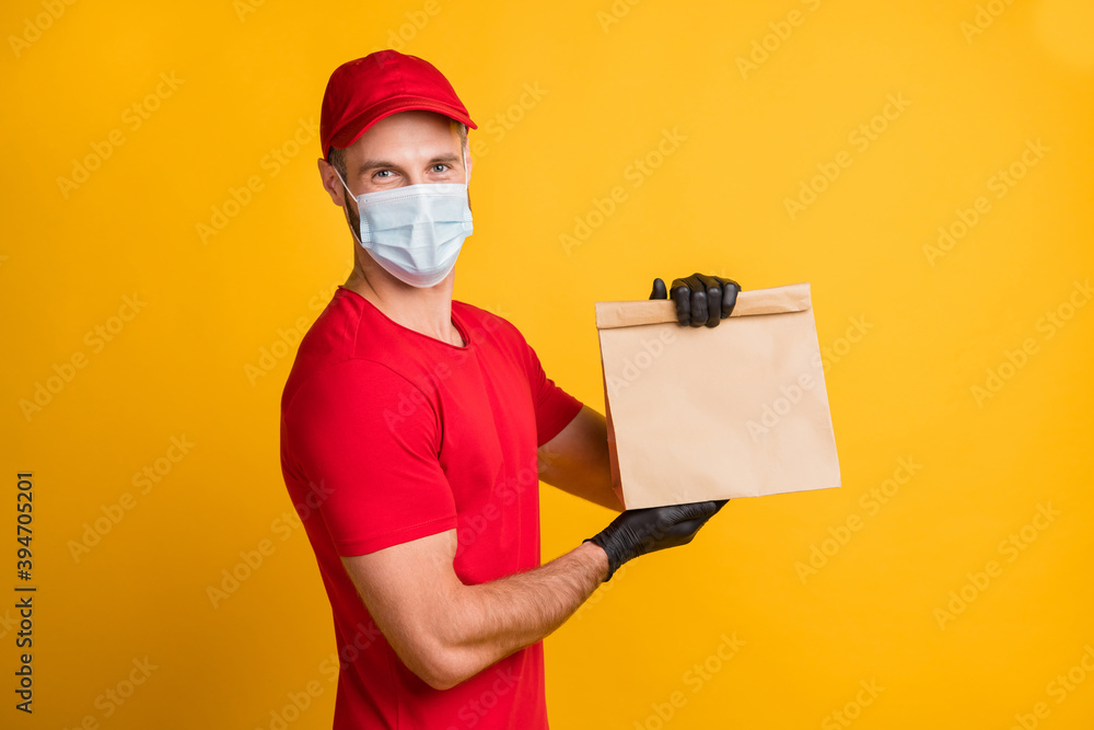 Photo of delivery guy hold takeaway package look camera wear mask red t-shirt cap isolated yellow color background Photo of delivery guy hold takeaway package look camera wear mask red t-shirt cap isolated yellow color background