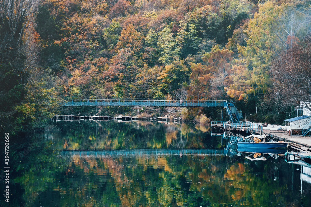 Suspension bridge with reflection in river or lake water, against mountain covered with yellow-green autumn forest with trees and small pier with boat in foreground Suspension bridge with reflection in river or lake water, against mountain covered with yellow-green autumn forest with trees and small pier with boat in foreground