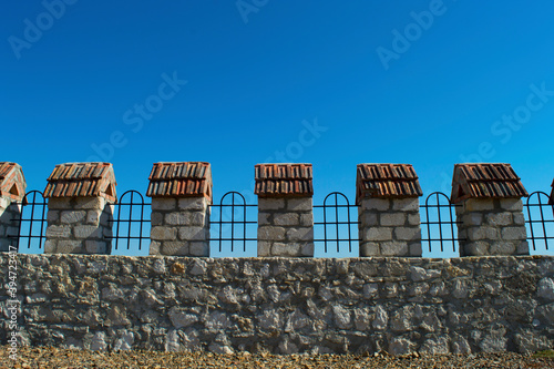 Rough old gray stone fence with tiled trellises and posts. The battlements of the fortress wall against the blue sky. Construction, design. Copy space.