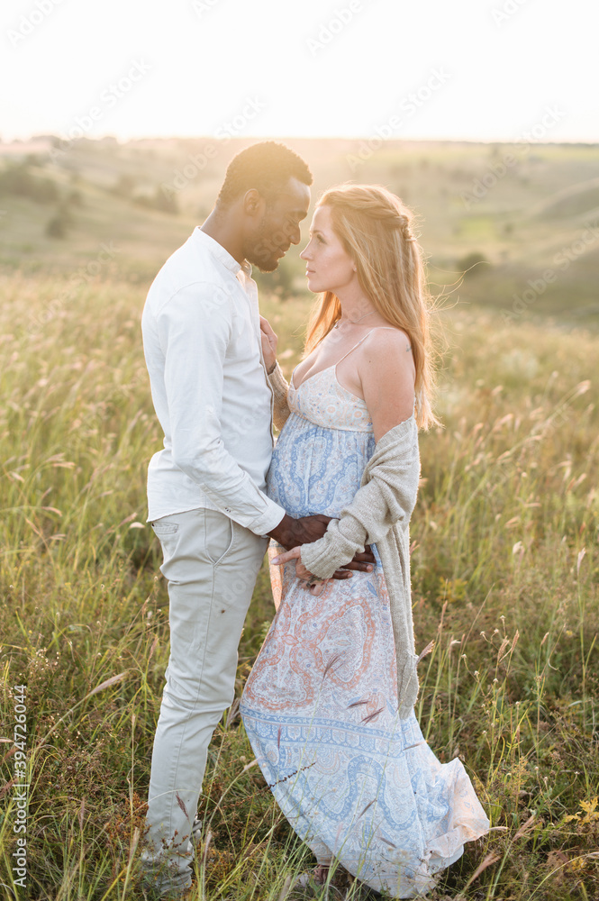 Stylish diverse couple. An african-american husband and his caucasian pregnant wife are holding hands and hugging outdoors Stylish diverse couple. An african-american husband and his caucasian pregnant wife are holding hands and hugging outdoors