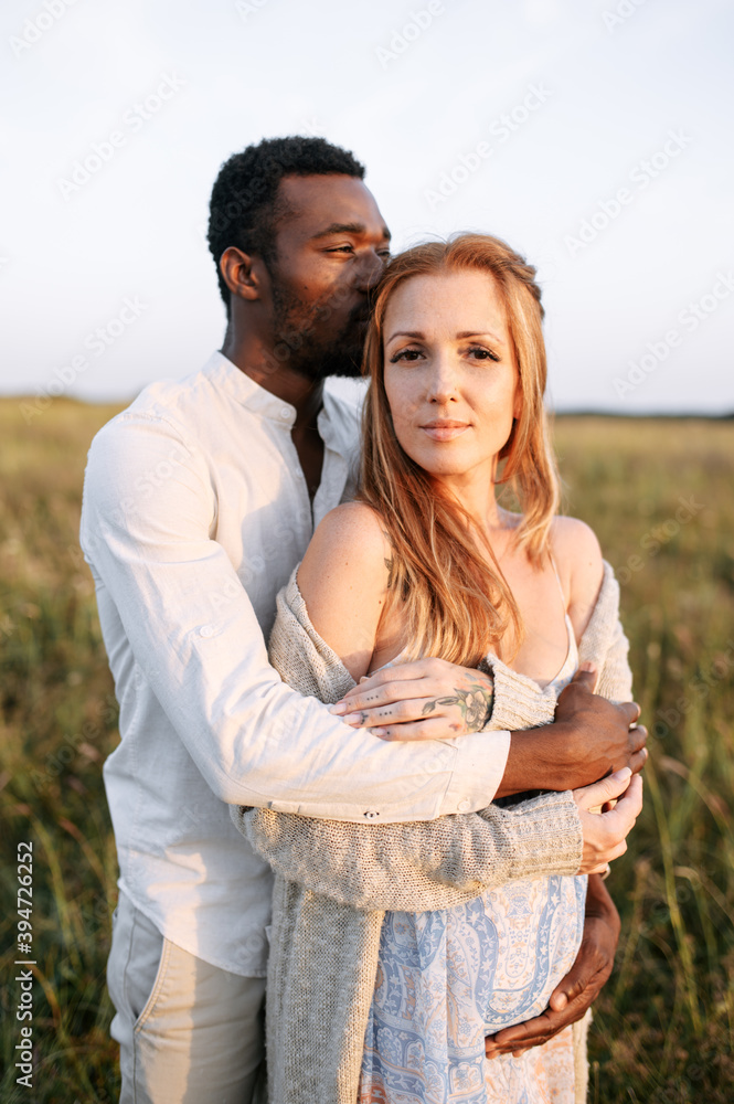 Obraz premium Summer love story on the meadow. An african american man in hugging charming pregnant young woman with care and awe
