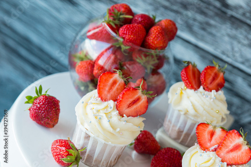 cupcake in the center with white cream decorated with ripe strawberries on a gray blue background and scattered strawberries.
