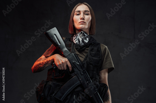 Weared with dark protective clothing female mercenary with short haircut and tattooed hand holding ak74 rifle in dark background.