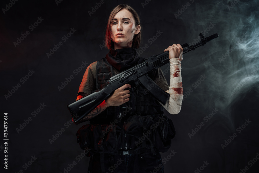 Dressed in dark armour military woman with short haircut and bandaged hand holds her assault rifle in dark smokey background. Dressed in dark armour military woman with short haircut and bandaged hand holds her assault rifle in dark smokey background.