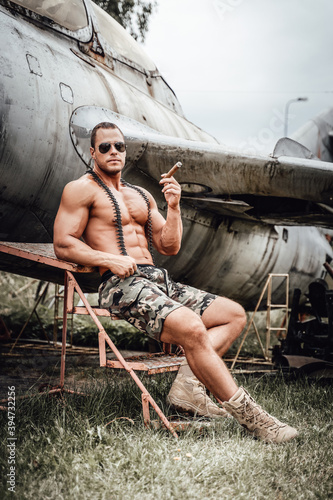 Sitting near abandoned airplane american soldier with naked torso and muscular build weared with sunglasses and smoking cigar.
