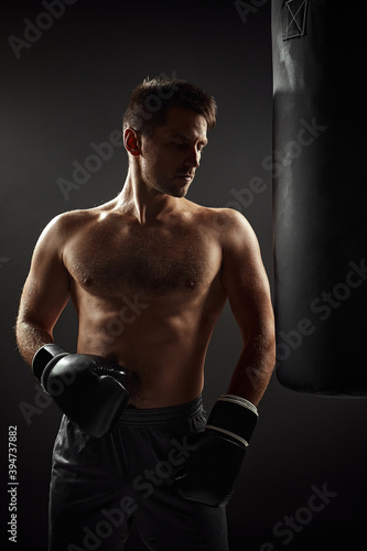 Young handsome male boxer in black gloves standing near punching bag on dark background