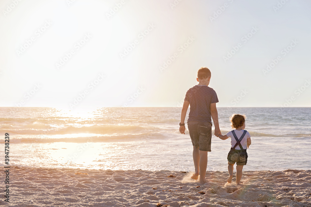 Boy And Little Girl Walking Towards The Ocean At Sunset Boy And Little Girl Walking Towards The Ocean At Sunset