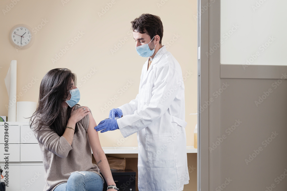 White woman with long hair wearing protective mask looking to the doctor speaking with him while being vaccinated on a clinic.  Vaccine for the pandemics concept with door frame negative space.