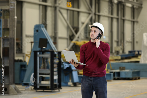 Portrait of a male factory manager in a white hard hat and red sweater holding laptop and mobile phone. Controlling the work process in the helicopter manufacturer. 