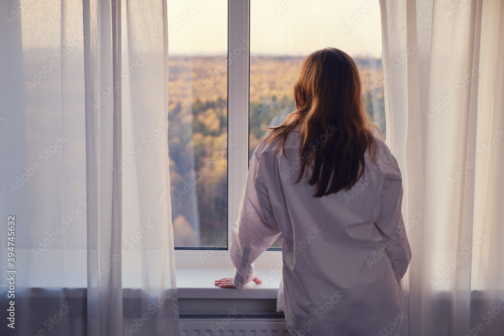 Redhead woman in white shirt by the window looking at autumn nature. Sad autumn weather and home comfort Redhead woman in white shirt by the window looking at autumn nature. Sad autumn weather and home comfort