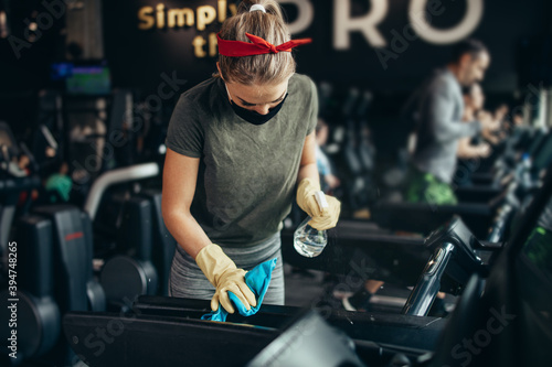 Young female worker disinfecting cleaning and weeping expensive fitness gym equipment with alcohol sprayer and cloth. Coronavirus global world pandemic and health protection safety measures.