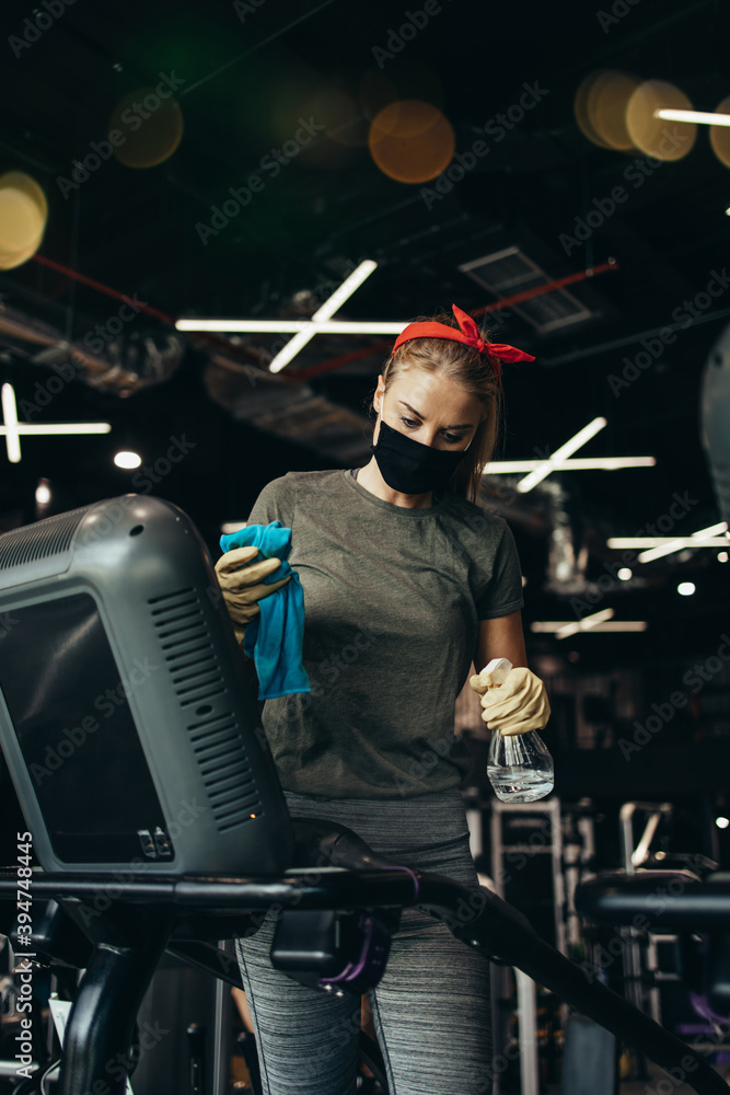 Fototapeta premium Young female worker disinfecting cleaning and weeping expensive fitness gym equipment with alcohol sprayer and cloth. Coronavirus global world pandemic and health protection safety measures.