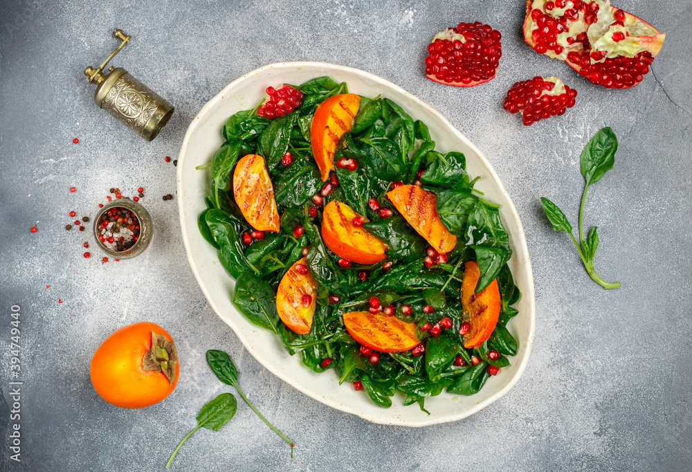 Φωτογραφία Delicious spinach salad with fried persimmon, pomegranate with olive oil dressing and spices in a white plate on a gray concrete or stone background