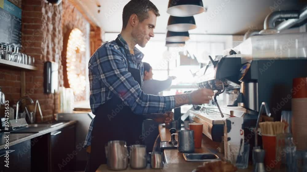 Handsome Male Barista in Checkered Shirt is Making a Take Away Cappuccino in Disposable Recycle Cup for a Customer in a Coffee Shop Bar. Female Cashier Works at a Cozy Cafe Counter in the Background. Handsome Male Barista in Checkered Shirt is Making a Take Away Cappuccino in Disposable Recycle Cup for a Customer in a Coffee Shop Bar. Female Cashier Works at a Cozy Cafe Counter in the Background.