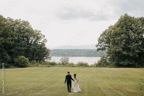 Fototapeta Naklejka Na Ścianę i Meble -  bride and groom walk toward water in the middle of a meadow on cloudy day