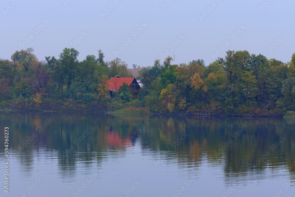 Astonishing landscape of autumn colored trees are reflected in the water. Wooden house with red tile roof is hidden in the forest near the river bank. Foggy autumn morning. Kyiv, Ukraine