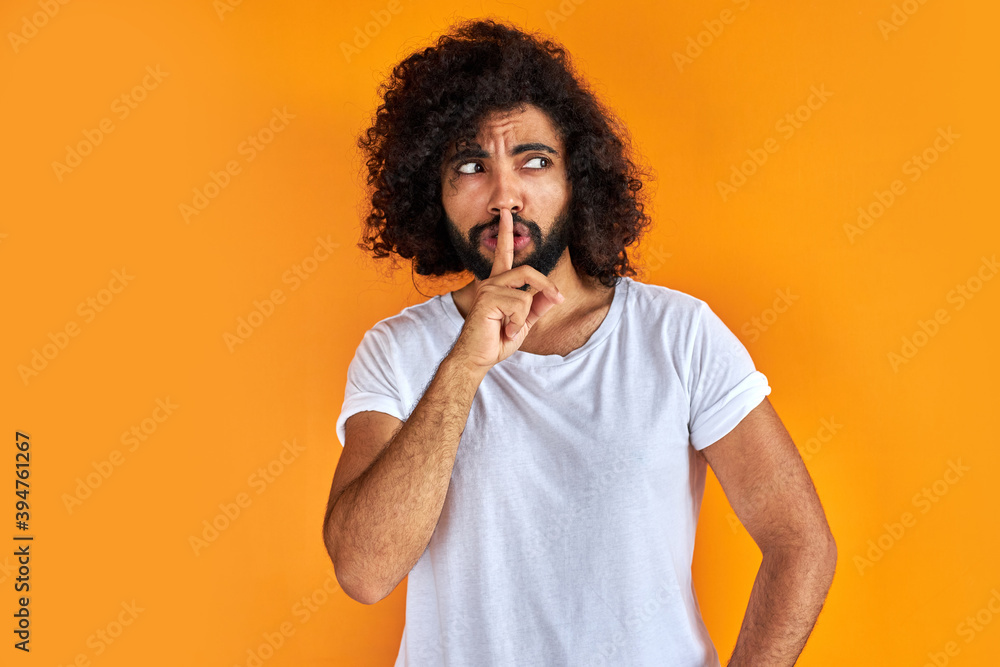 young arab man with curly black hair raising his finger on the front of his mouth asking to be quiet, isolated on orange background, he is looking away young arab man with curly black hair raising his finger on the front of his mouth asking to be quiet, isolated on orange background, he is looking away