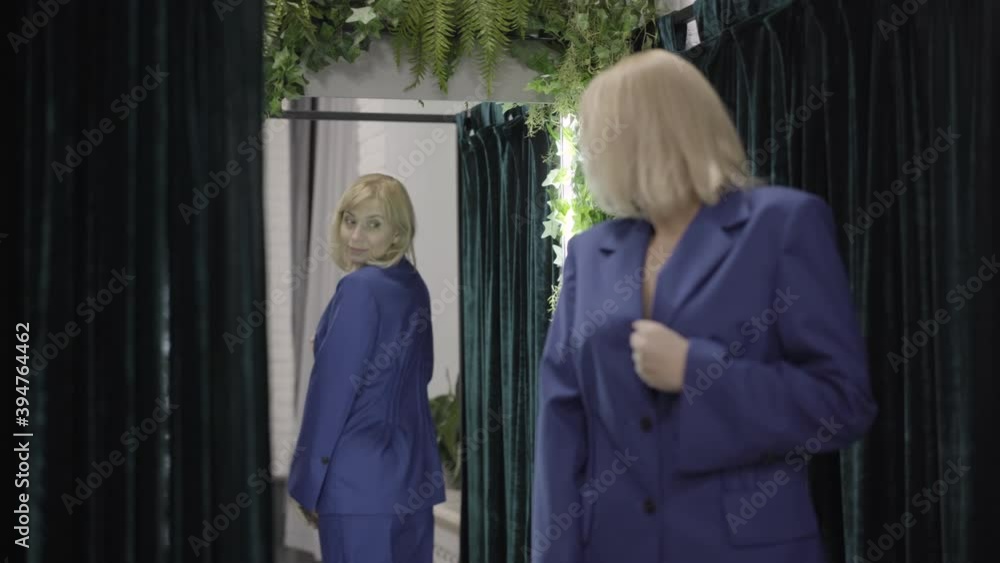 Back view of confident elegant Caucasian woman trying on clothes in shop dressing room. Rack focus changes from shopper to reflection in mirror. Shopping and fashion concept. Back view of confident elegant Caucasian woman trying on clothes in shop dressing room. Rack focus changes from shopper to reflection in mirror. Shopping and fashion concept.