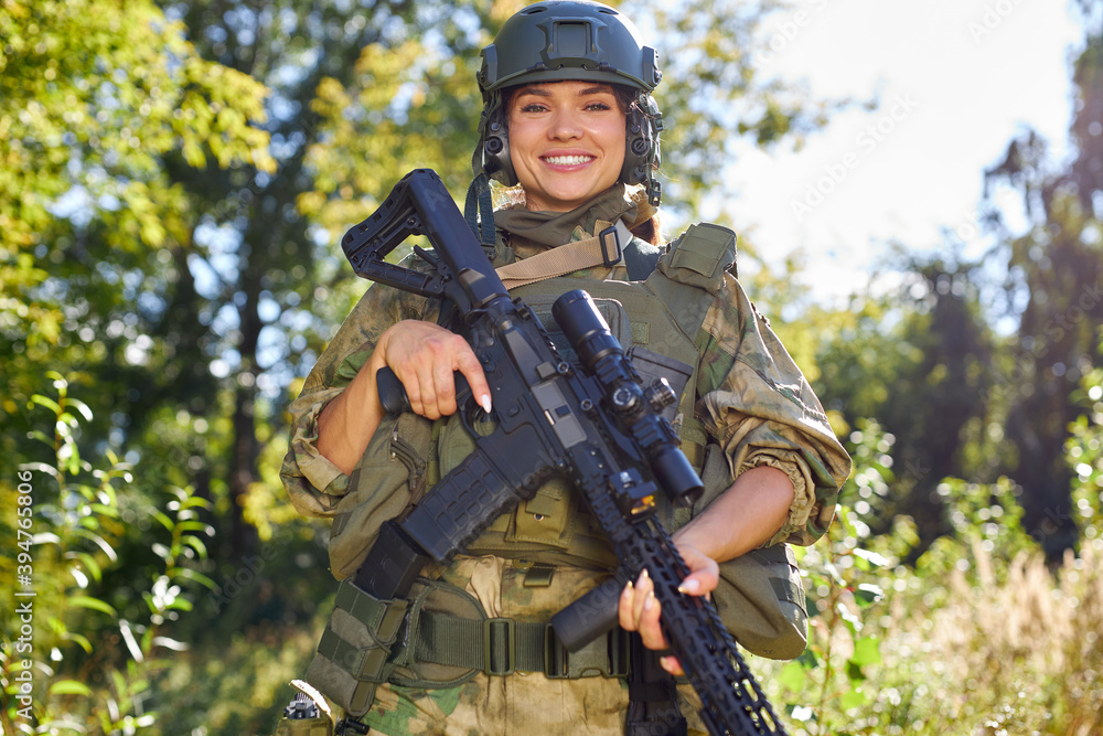 portrait of cheerful smiling female soldier with rifle gun in hands in green military suit and hat, in wild nature forest