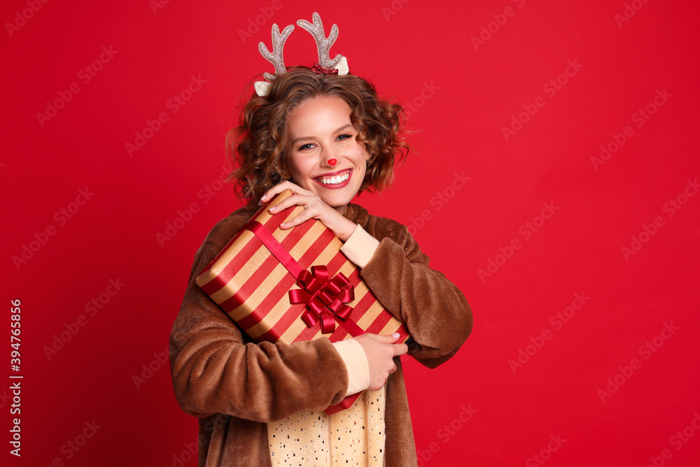 cute funny young woman in a Christmas reindeer costume smiles and rejoices with gift on colorful red background. cute funny young woman in a Christmas reindeer costume smiles and rejoices with gift on colorful red background.
