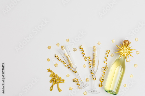 Festive white background with gold decoration , bottle of sparkling wine with two crystal glasses, shiny golden serpentine confetti and glittering snowflakes, golden fir-tree, copy space, top view