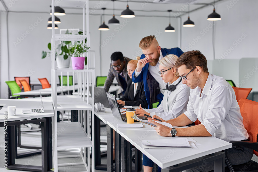 male director give directions to employees in the office, young team sit working together, using laptop and papers male director give directions to employees in the office, young team sit working together, using laptop and papers