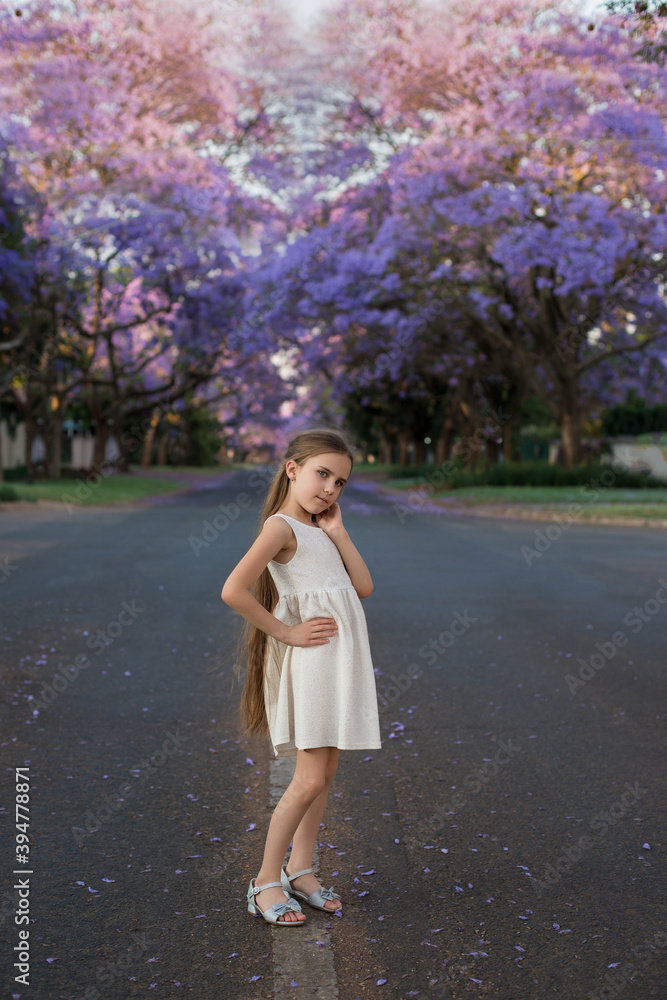A little cute girl with long hair in a white dress stands on the street with blooming jacaranda trees.