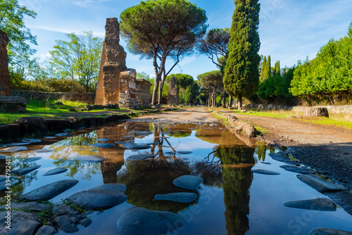 Appia Antica, Rome, reflects the remains of the graves in the puddle, after the storm with the blue sky and clouds on an autumn day. Regina Viarum, Italy.