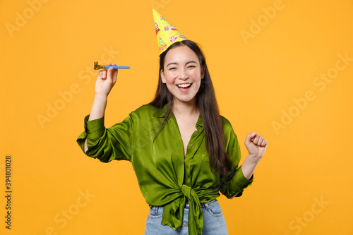 Photography Funny cheerful beautiful young brunette asian woman wearing basic green shirt birthday hat hold pipe dancing clenching fist looking camera isolated on bright yellow colour background, studio portrait