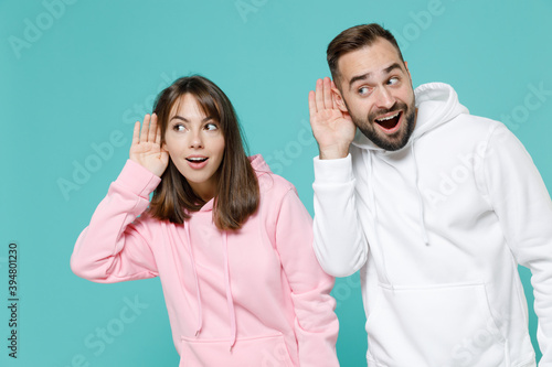 Curious shocked young couple two friends man woman 20s wearing white pink casual hoodie try to hear you overhear listening intently isolated on blue turquoise colour wall background studio portrait.