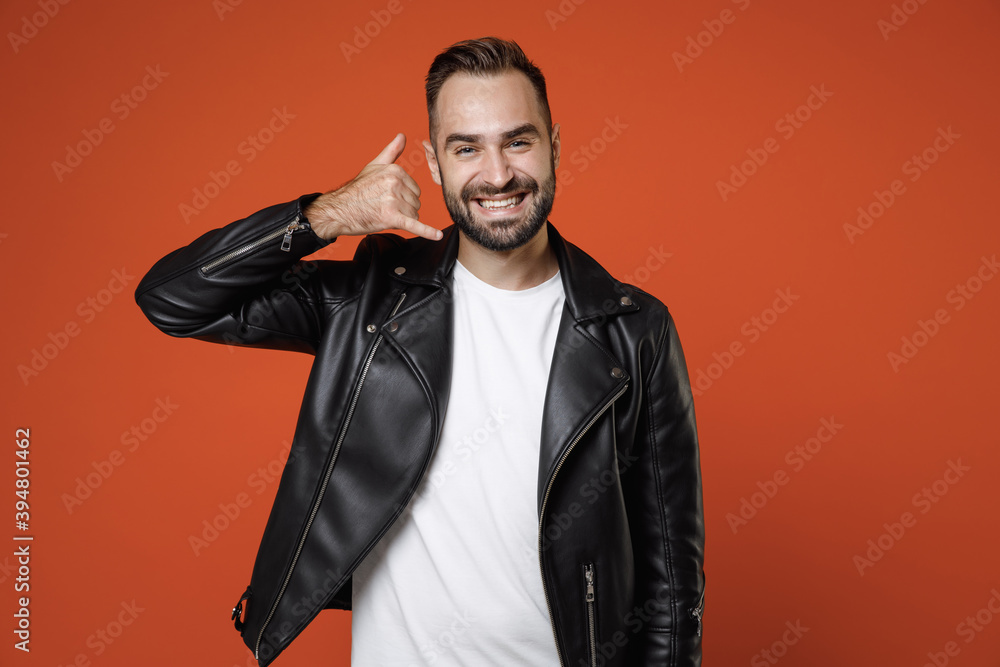 Smiling young bearded man in basic white t-shirt black leather jacket standing doing phone gesture like says call me back looking camera isolated on bright orange colour background studio portrait. Smiling young bearded man in basic white t-shirt black leather jacket standing doing phone gesture like says call me back looking camera isolated on bright orange colour background studio portrait.