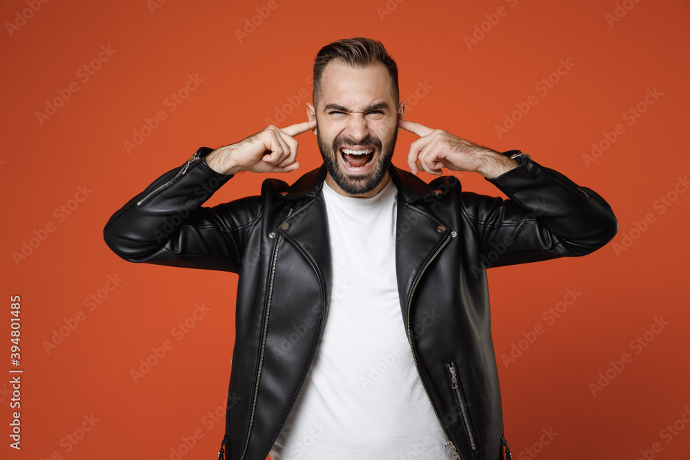 Frustrated young bearded man in basic white t-shirt black leather jacket standing covering ears with fingers looking camera screaming isolated on bright orange colour wall background studio portrait. Frustrated young bearded man in basic white t-shirt black leather jacket standing covering ears with fingers looking camera screaming isolated on bright orange colour wall background studio portrait.