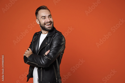 Side view of smiling cheerful young bearded man 20s wearing basic white t-shirt black leather jacket standing holding hands crossed looking aside isolated on orange colour background studio portrait.