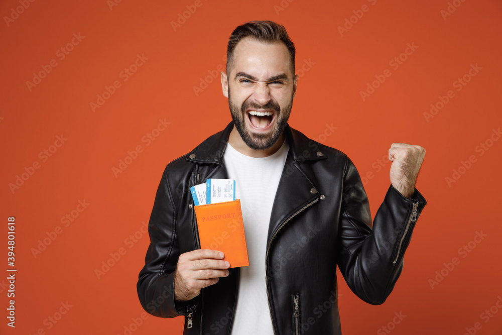 Happy young traveler tourist man in white t-shirt, leather jacket hold passport tickets doing winner gesture isolated on orange background. Passenger traveling on weekends. Air flight journey concept. Happy young traveler tourist man in white t-shirt, leather jacket hold passport tickets doing winner gesture isolated on orange background. Passenger traveling on weekends. Air flight journey concept.