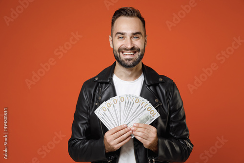 Obraz na plátně Smiling young bearded man in basic white t-shirt, black leather jacket standing hold fan of cash money in dollar banknotes looking camera isolated on bright orange colour background studio portrait