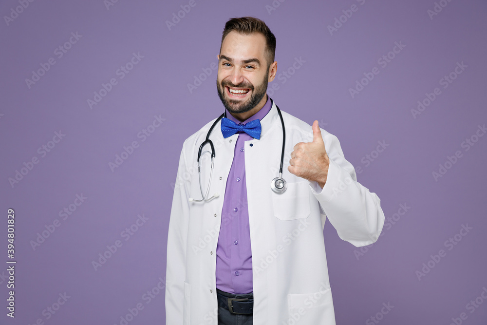 Smiling cheerful young bearded doctor man wearing white medical gown showing thumb up looking camera isolated on violet colour background studio portrait. Healthcare personnel health medicine concept. Smiling cheerful young bearded doctor man wearing white medical gown showing thumb up looking camera isolated on violet colour background studio portrait. Healthcare personnel health medicine concept.