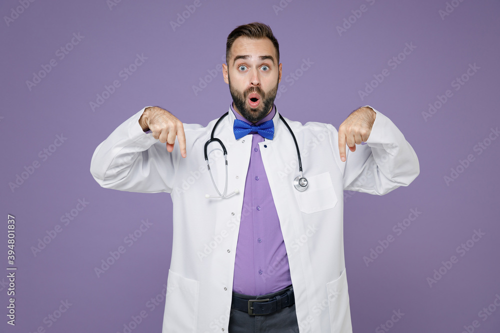 Shocked young bearded doctor man in white medical gown pointing index fingers down on mock up copy space isolated on violet background studio portrait. Healthcare personnel health medicine concept. Shocked young bearded doctor man in white medical gown pointing index fingers down on mock up copy space isolated on violet background studio portrait. Healthcare personnel health medicine concept.