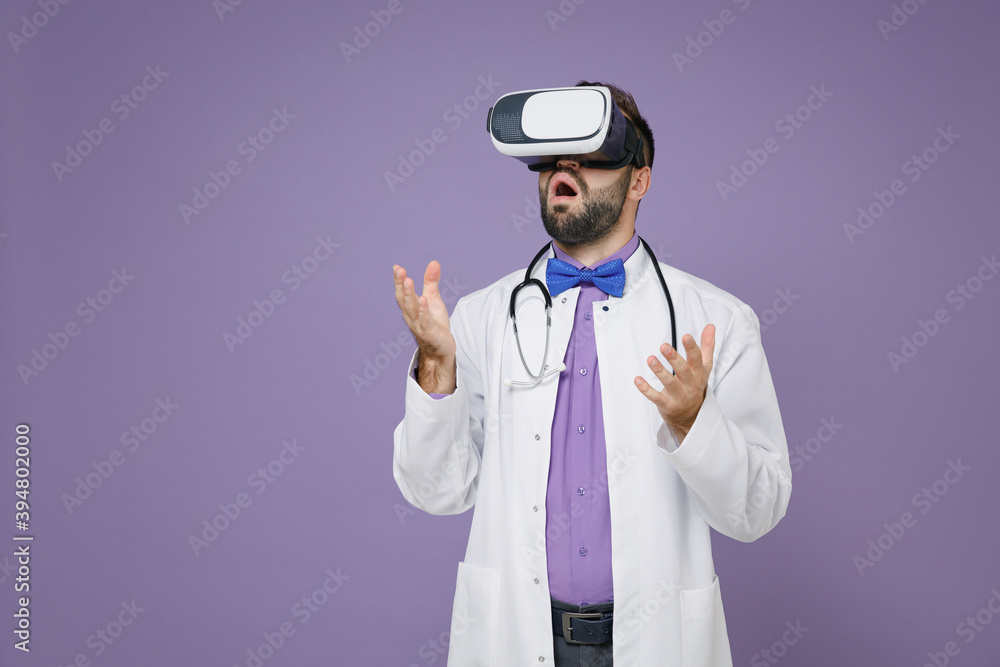 Shocked young bearded doctor man in white medical gown stethoscope watching in vr headset gadget spreading hands isolated on violet background in studio. Healthcare personnel health medicine concept. Shocked young bearded doctor man in white medical gown stethoscope watching in vr headset gadget spreading hands isolated on violet background in studio. Healthcare personnel health medicine concept.