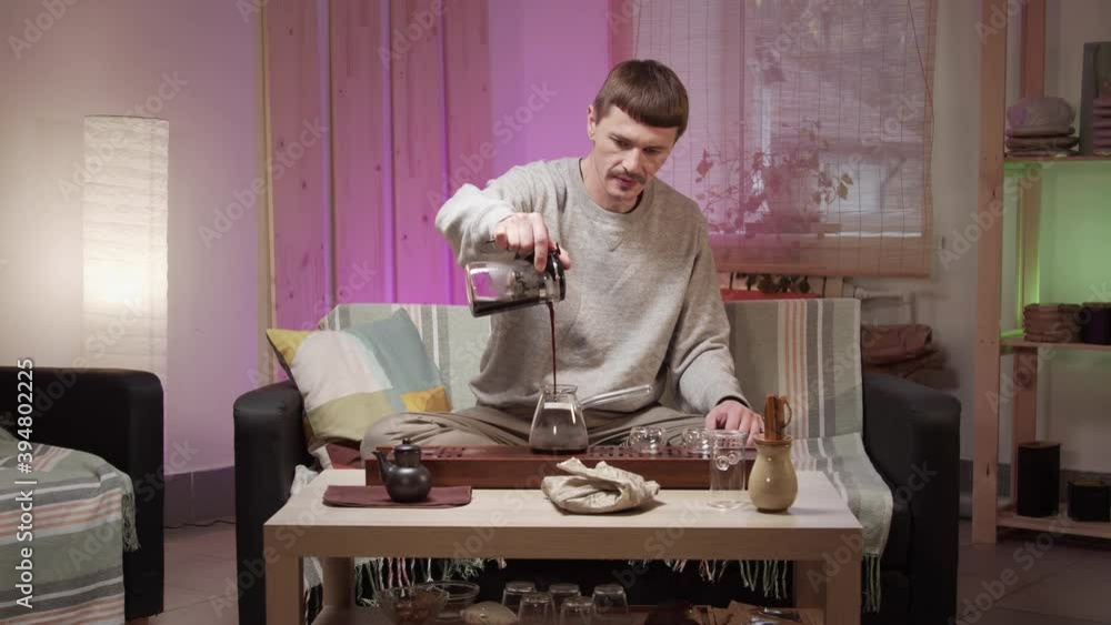 A middle aged man holding a teapot of brewed tea high pours the drink into a glass cup and drinks the tea A middle aged man holding a teapot of brewed tea high pours the drink into a glass cup and drinks the tea