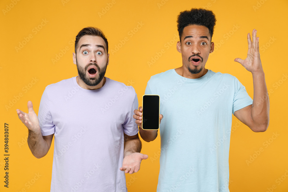 Shocked young two friends european african american men 20s wearing violet blue t-shirts using mobile cell phone with blank empty screen spreading hands isolated on yellow background studio portrait. Shocked young two friends european african american men 20s wearing violet blue t-shirts using mobile cell phone with blank empty screen spreading hands isolated on yellow background studio portrait.
