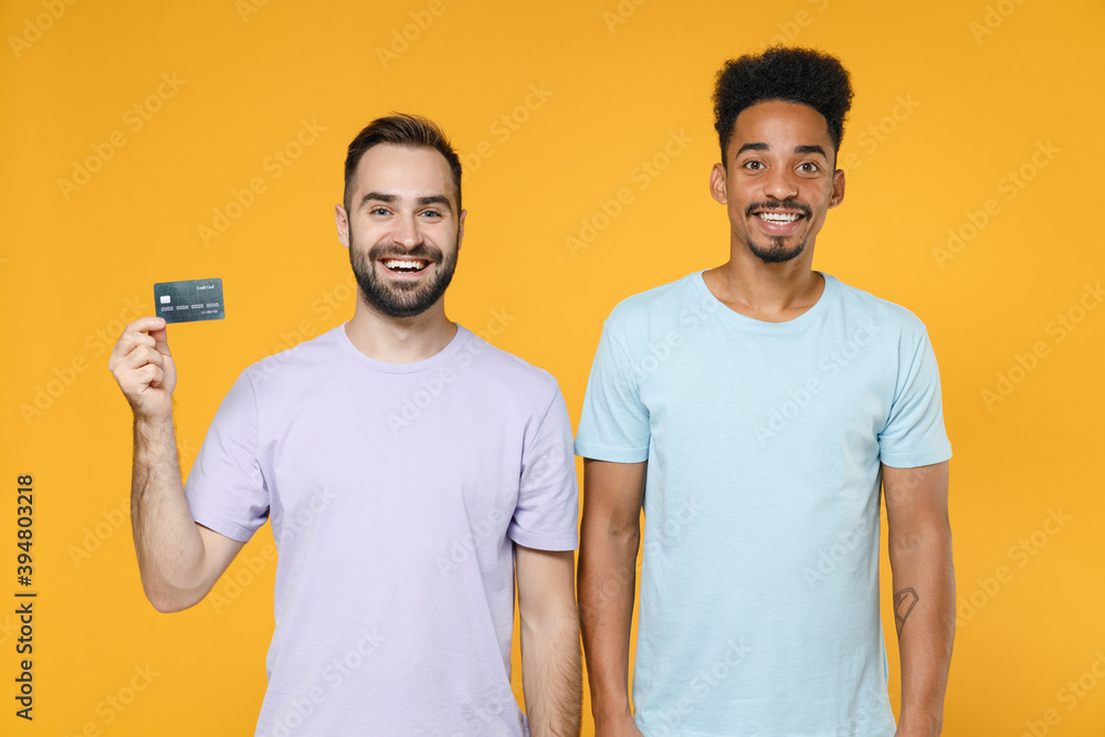 Cheerful young two friends european african american men 20s wearing violet blue casual t-shirts hold credit bank card looking camera isolated on bright yellow colour wall background studio portrait. Cheerful young two friends european african american men 20s wearing violet blue casual t-shirts hold credit bank card looking camera isolated on bright yellow colour wall background studio portrait.
