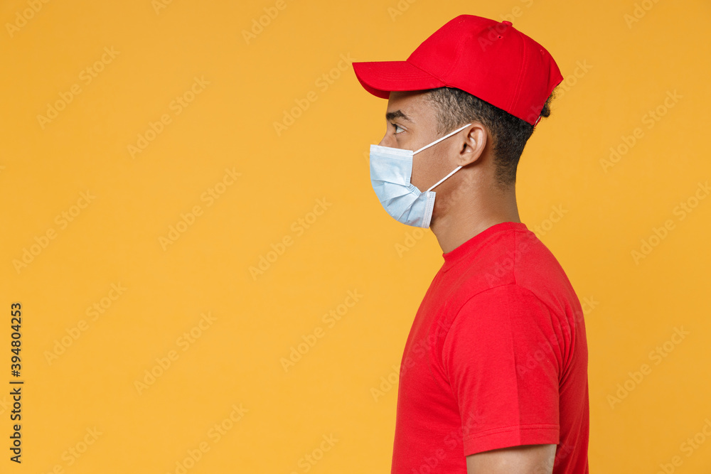 Side view delivery employee african man in red cap blank print tshirt face mask gloves work courier dealer service on quarantine coronavirus covid19 virus concept isolated on yellow background studio Side view delivery employee african man in red cap blank print tshirt face mask gloves work courier dealer service on quarantine coronavirus covid19 virus concept isolated on yellow background studio