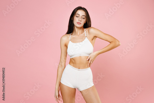 Charming tender beautiful young brunette woman 20s in white underwear demonstrating perfect fit body standing posing put hand on waist isolated on pastel pink colour background, studio portrait.