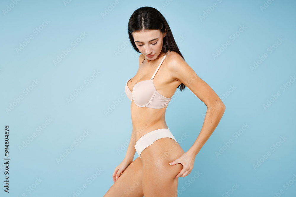 Side view of beautiful young brunette woman 20s in beige underwear with sports fit body standing posing checking cellulite on her booty isolated on pastel blue colour wall background studio portrait. Side view of beautiful young brunette woman 20s in beige underwear with sports fit body standing posing checking cellulite on her booty isolated on pastel blue colour wall background studio portrait.