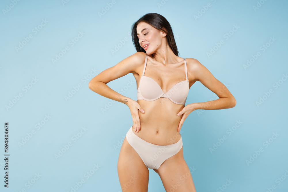 Smiling charming beautiful cute young brunette woman 20s in beige underwear showing fit body standing posing hold hands on waist looking aside isolated on pastel blue background, studio portrait. Smiling charming beautiful cute young brunette woman 20s in beige underwear showing fit body standing posing hold hands on waist looking aside isolated on pastel blue background, studio portrait.