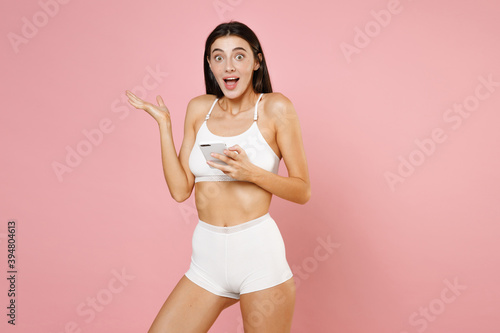 Excited surprised amazed young brunette woman 20s wearing white underwear posing using mobile cell phone typing sms message spreading hands isolated on pastel pink colour background studio portrait.